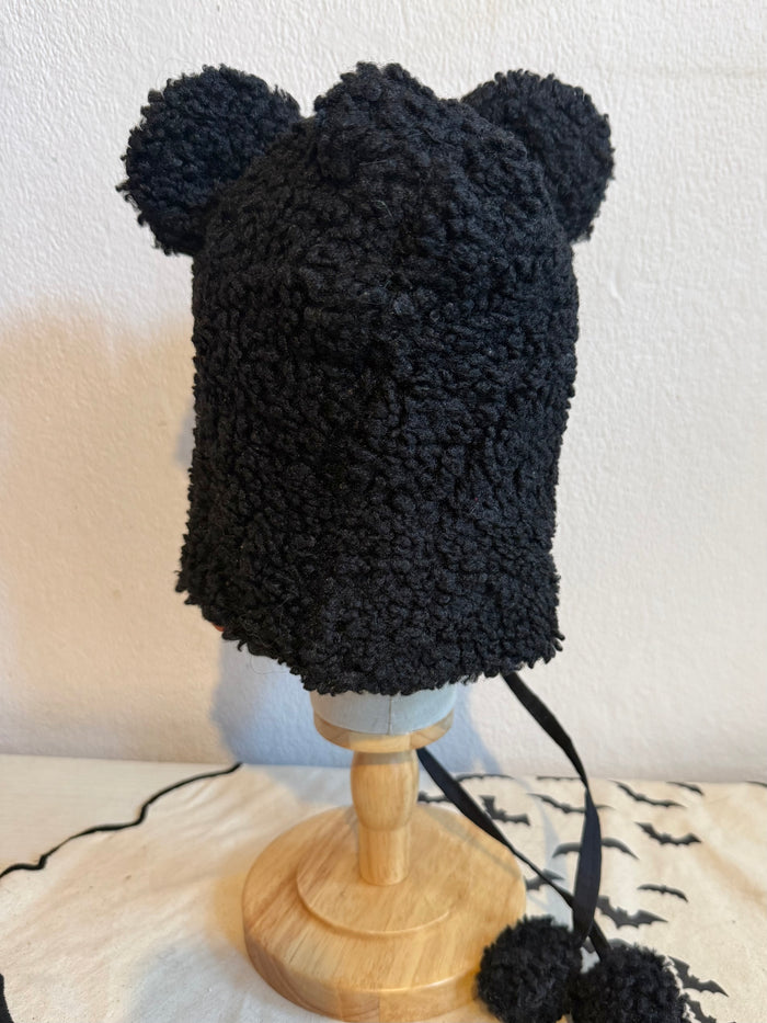 Black fuzzy bear-ear bonnet with a black ribbon bow and pom-poms, displayed on a mannequin bust against a beige wall.