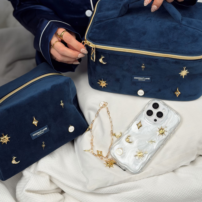 Woman wearing satin navy pyjamas with a phone in a stardust after dark phone case with gold charm embellishments. The phone has a matching gold chain phone strap with celestial style charms, including a moon, sun, star, diamond shape and 11:11 charm.
In the photo is the matching makeup bag and vanity case with navy velvet material and gold charm embellishments.
