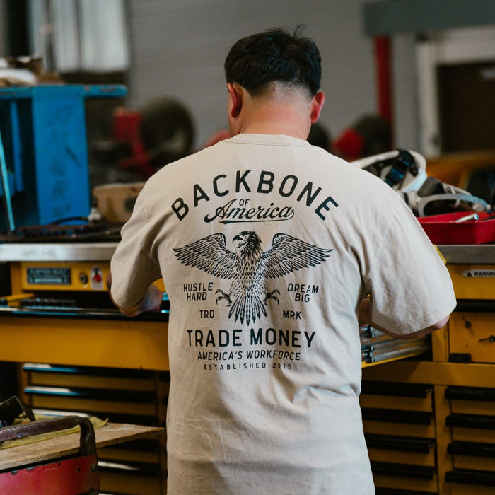 Trade Money America's Workforce graphic t-shirt in faded khaki, shown worn in a workshop setting