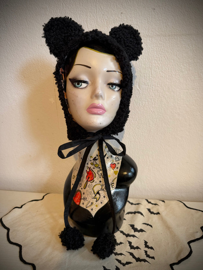 Black fuzzy bear-ear bonnet with a black ribbon bow and pom-poms, displayed on a mannequin bust against a beige wall.