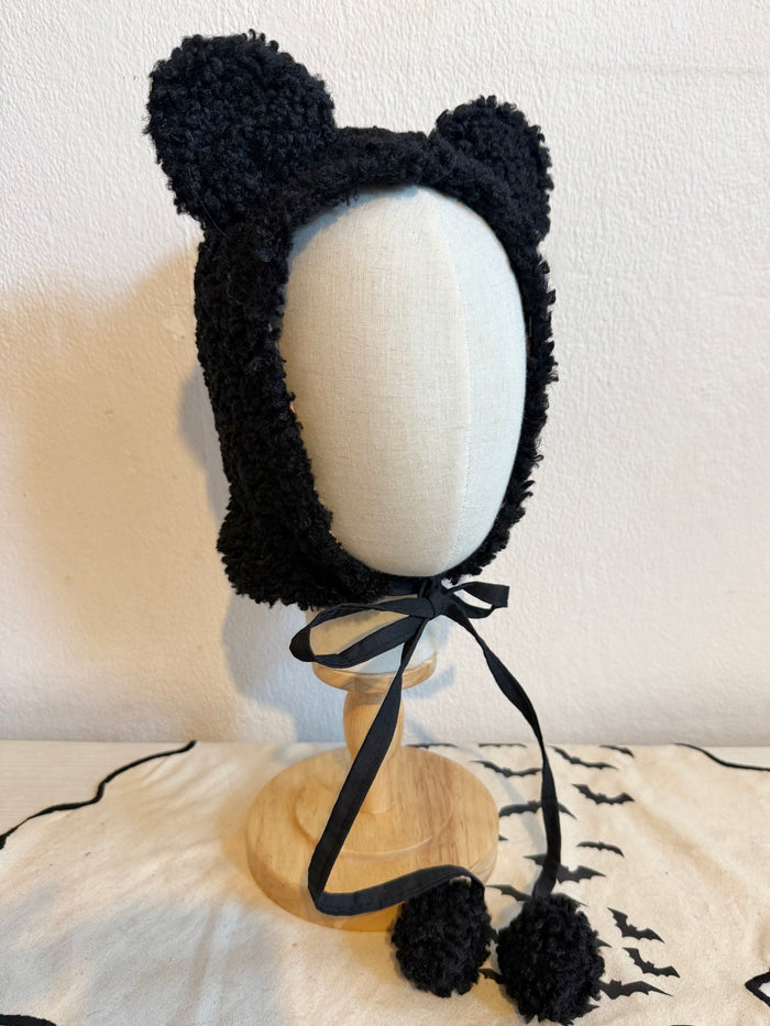 Black fuzzy bear-ear bonnet with a black ribbon bow and pom-poms, displayed on a mannequin bust against a beige wall.