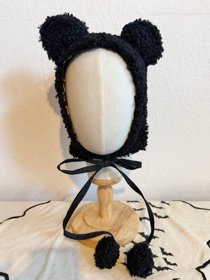 Black fuzzy bear-ear bonnet with a black ribbon bow and pom-poms, displayed on a mannequin bust against a beige wall.