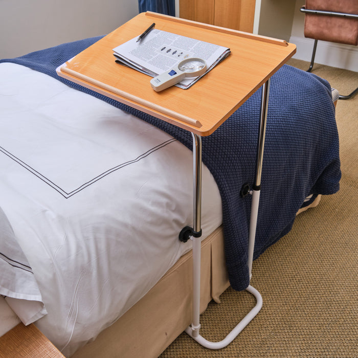 Angled view of NRS Divan Overbed Table with beech-effect tilting table top and white U-shaped base on castors against a neutral background.