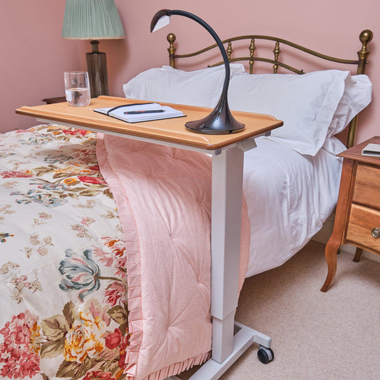 Angled view of NRS Easylift Overbed Table with beech-effect tilting table top and white U-shaped base on castors against a neutral background.