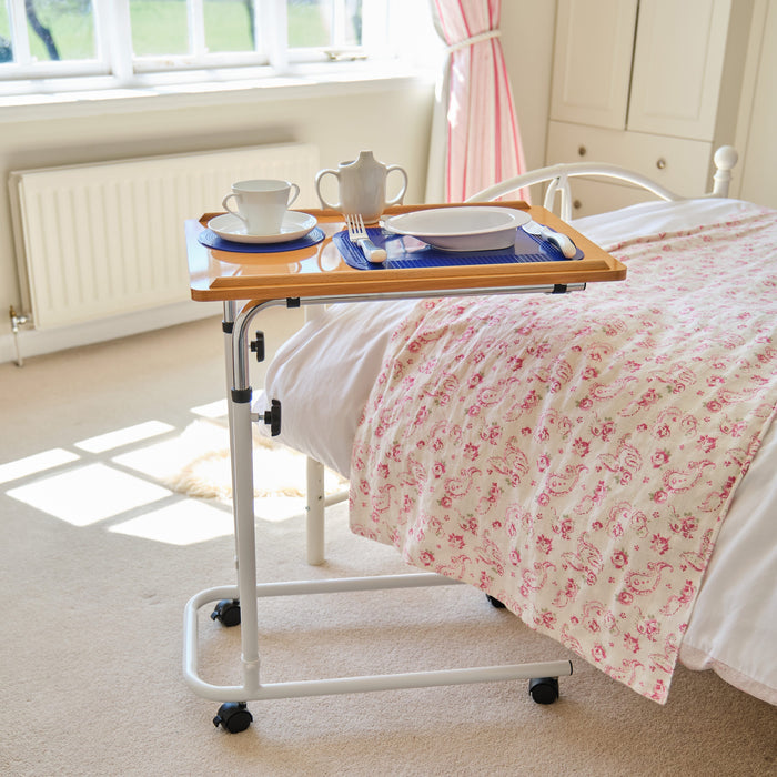 Angled view of NRS Overbed Table with beech-effect tilting table top and white U-shaped base on castors against a neutral background.