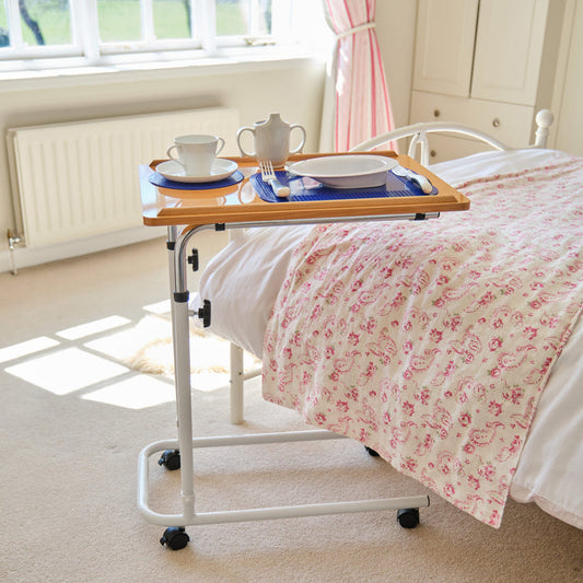 Angled view of NRS Overbed Table with beech-effect tilting table top and white U-shaped base on castors against a neutral background.
